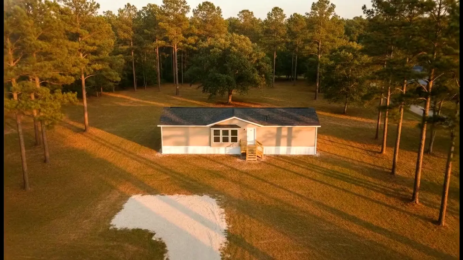 Aerial view of a double-wide mobile home set up on its own land in the East Texas pines at sunset, with skirting installed, wooden entry steps and a gravel drive